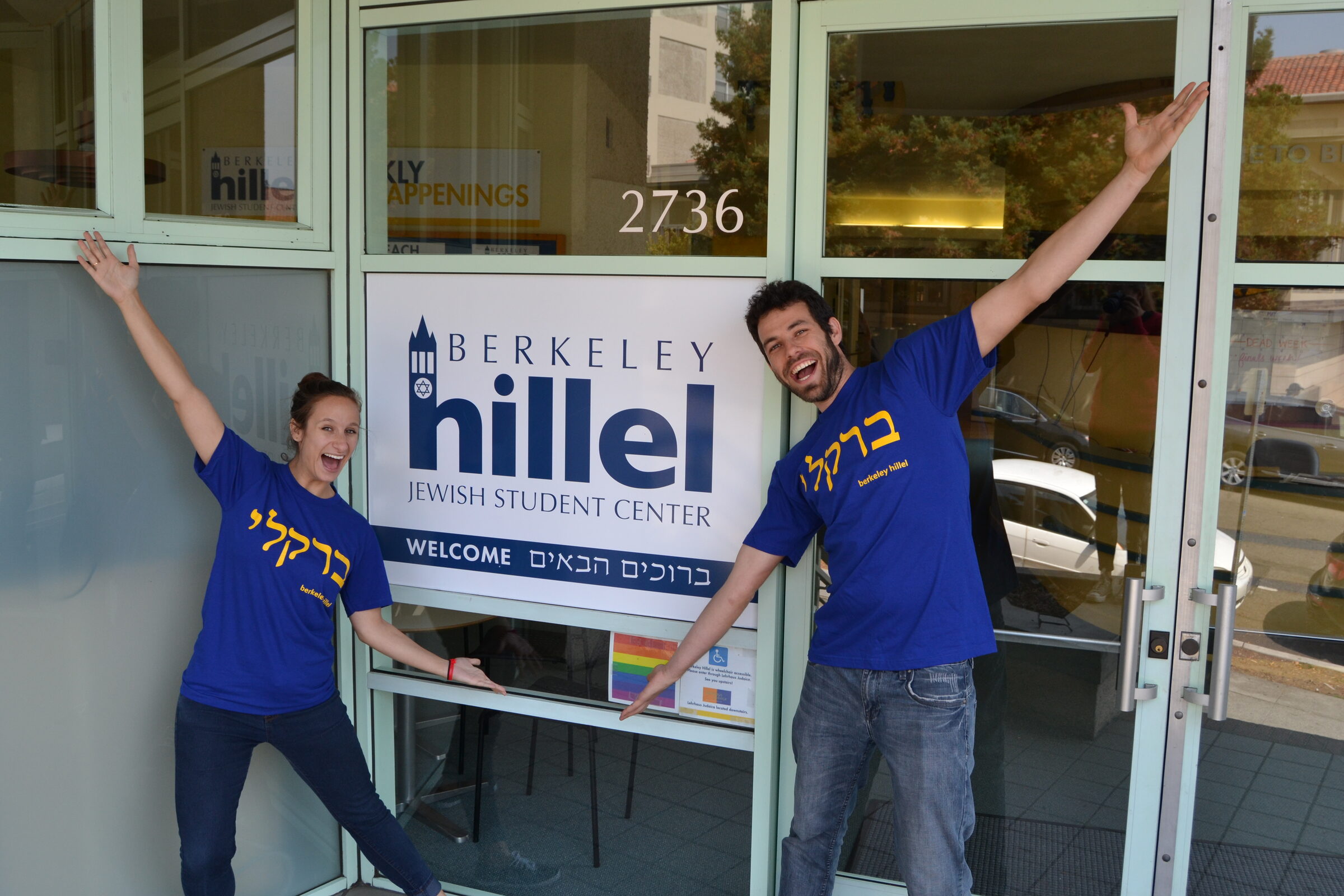 Two staff members in blue Berkeley Hillel shirts throwing their arms wide at the entrance to 2736 Bancroft Way